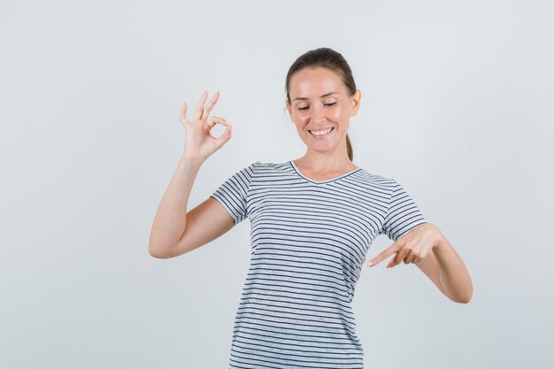 Mujer joven apuntando hacia abajo con signo ok en camiseta y mirando contento. vista frontal.