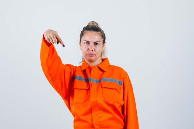 Mujer joven apuntando hacia abajo en uniforme de trabajador y mirando serio, vista frontal.