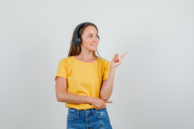 Mujer joven apuntando hacia afuera en camiseta, pantalones cortos, auriculares y mirando contento. vista frontal.