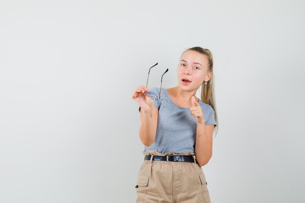 Mujer joven apuntando al frente mientras sostiene gafas en camiseta, vista frontal de pantalones.