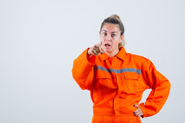 Mujer joven apuntando al frente con uniforme de trabajador y mirando nerviosa. vista frontal.