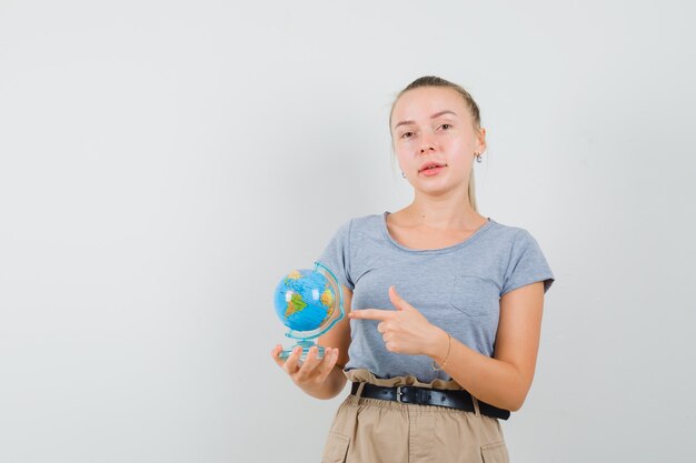 Mujer joven apuntando al globo terráqueo en camiseta, pantalón y mirando confiado. vista frontal.