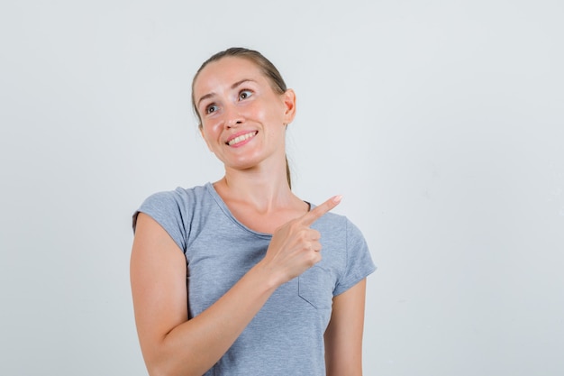 Mujer joven apuntando al lado en camiseta gris y mirando alegre, vista frontal.