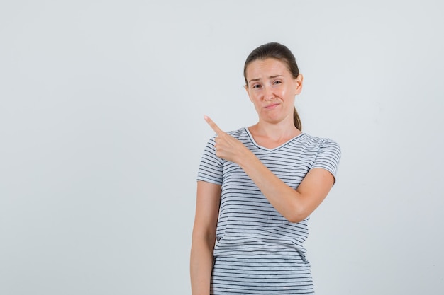 Mujer joven apuntando al lado en camiseta y mirando insatisfecho. vista frontal.