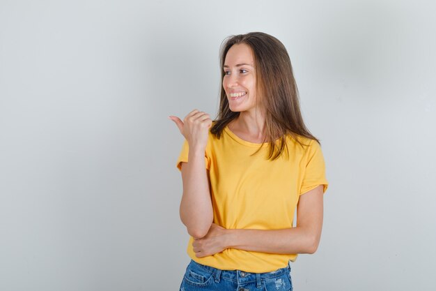 Mujer joven apuntando al lado con el pulgar y sonriendo en camiseta