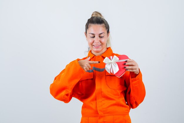 Mujer joven apuntando al regalo rojo en uniforme de trabajador y mirando complacido. vista frontal.