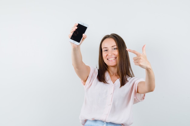 Mujer joven apuntando al teléfono móvil en camiseta, falda y mirando alegre. vista frontal.