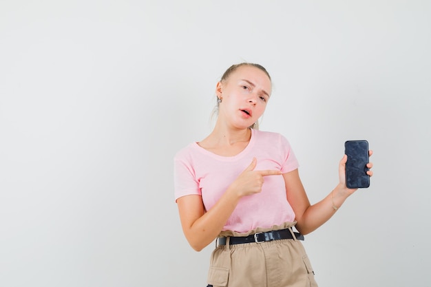 Mujer joven apuntando al teléfono móvil en camiseta, pantalón, vista frontal.