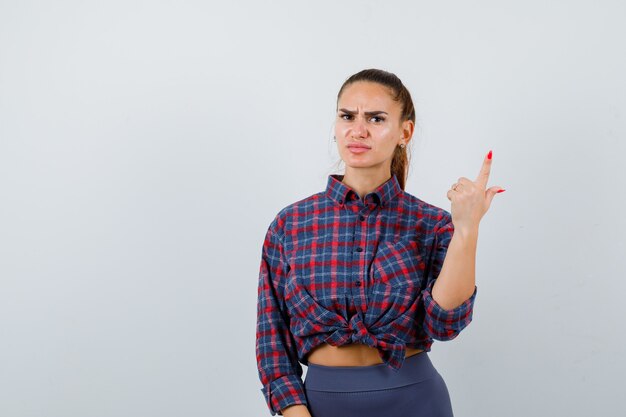 Mujer joven apuntando hacia arriba en camisa a cuadros, pantalones y mirando serio, vista frontal.