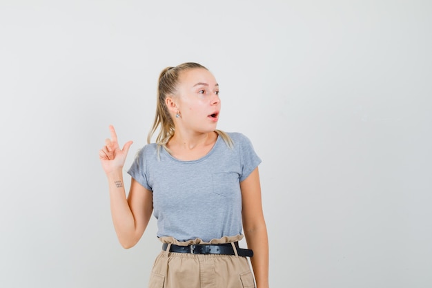 Mujer joven apuntando hacia arriba en camiseta, pantalón y mirando sorprendido. vista frontal.