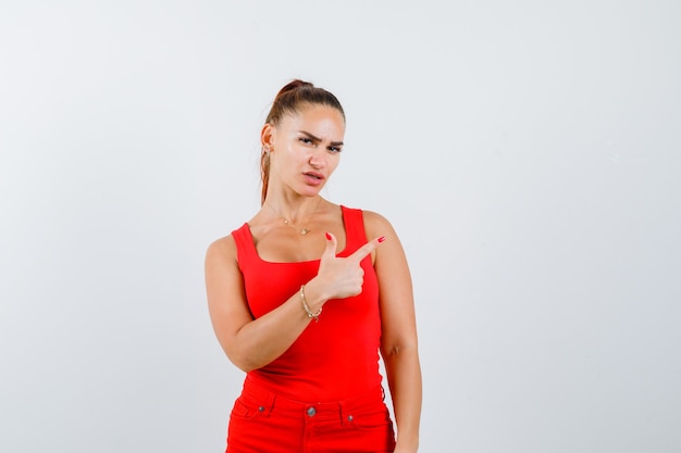 Mujer joven apuntando hacia arriba en camiseta roja, pantalones y mirando pensativo, vista frontal.