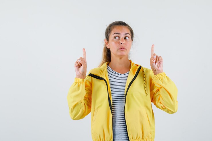 Foto gratuito mujer joven apuntando hacia arriba en chaqueta, camiseta y mirando pensativo, vista frontal.