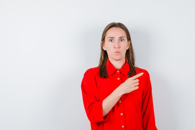 Mujer joven apuntando hacia arriba con el dedo en blusa roja y mirando sorprendido, vista frontal.