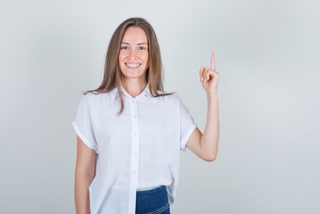 Mujer joven apuntando hacia arriba con el dedo y sonriendo en camiseta blanca