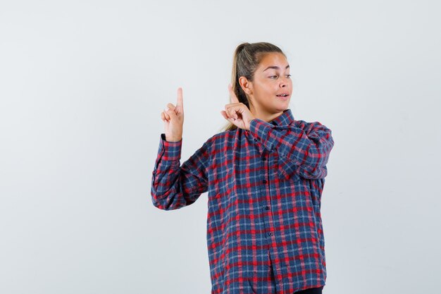 Foto gratuita mujer joven apuntando hacia arriba con los dedos índices en camisa a cuadros y mirando feliz