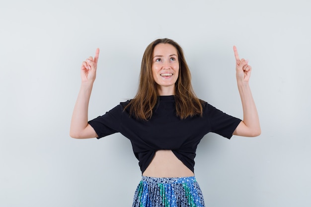Mujer joven apuntando hacia arriba con los dedos índices en camiseta negra y falda azul y mirando feliz