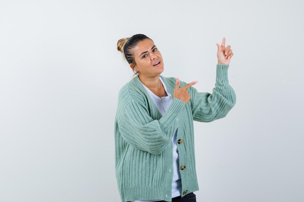 Foto gratuita mujer joven apuntando hacia arriba y hacia la derecha en camisa blanca y chaqueta de punto verde menta y mirando feliz