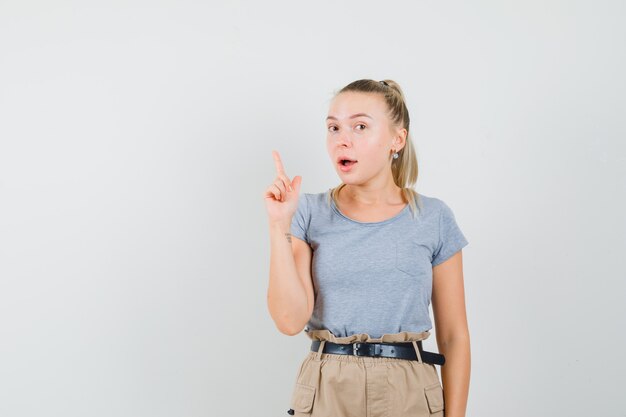 Mujer joven apuntando hacia arriba, con una excelente idea en camiseta, pantalón, vista frontal.