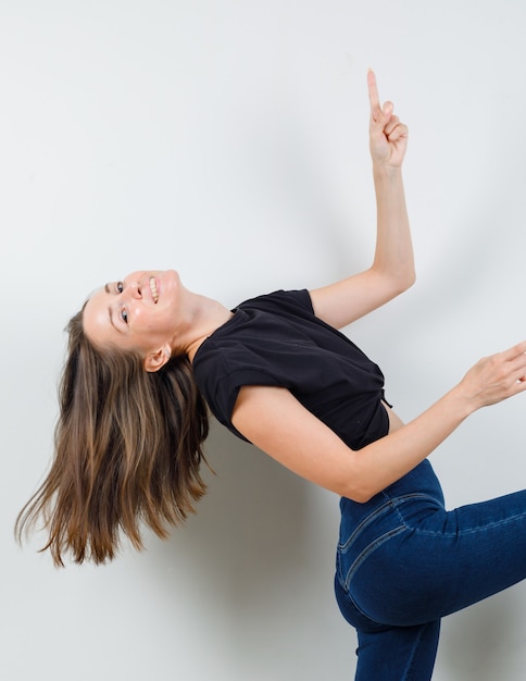 Foto gratuita mujer joven apuntando hacia arriba mientras levanta la pierna en blusa negra, pantalones y luciendo enérgico