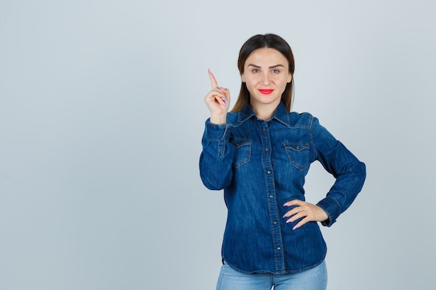 Mujer joven apuntando hacia arriba mientras mantiene la mano en la cadera en camisa de mezclilla y jeans y se ve alegre