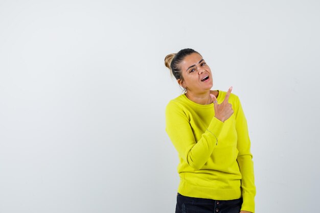 Mujer joven apuntando hacia arriba en suéter amarillo y pantalón negro y mirando feliz