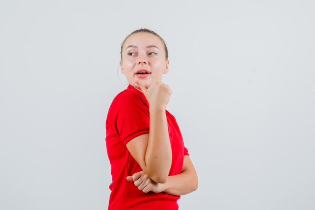 Mujer joven apuntando hacia atrás con el pulgar en camiseta roja y mirando curioso