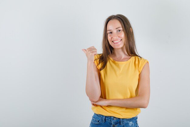 Mujer joven apuntando hacia atrás con su pulgar en camiseta, pantalones cortos y mirando alegre.