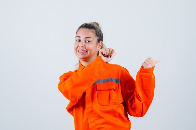 Mujer joven apuntando hacia atrás en uniforme de trabajador y mirando alegre, vista frontal.