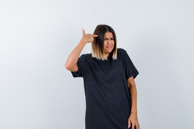 Mujer joven apuntando a la cabeza con vestido de polo y mirando disgustado. vista frontal.