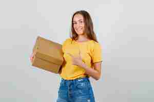 Foto gratuita mujer joven apuntando a la caja de cartón en camiseta, pantalones cortos y mirando contento. vista frontal.