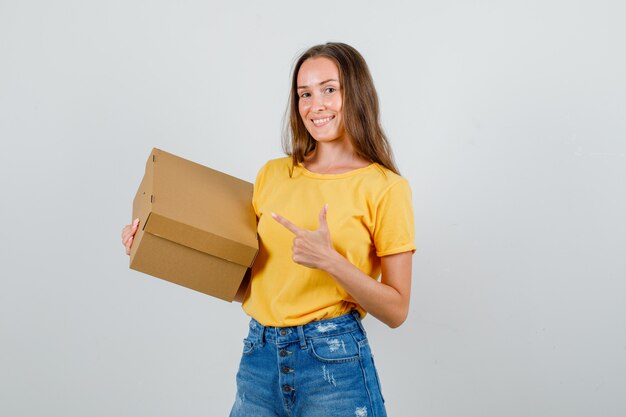 Mujer joven apuntando a la caja de cartón en camiseta, pantalones cortos y mirando contento. vista frontal.