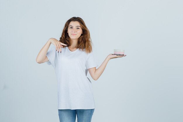 Mujer joven apuntando a caja de regalo en camiseta blanca, jeans y mirando contento. vista frontal.