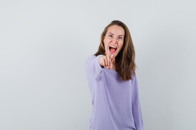 Mujer joven apuntando a la cámara mientras grita en blusa lila y parece agresiva. vista frontal.