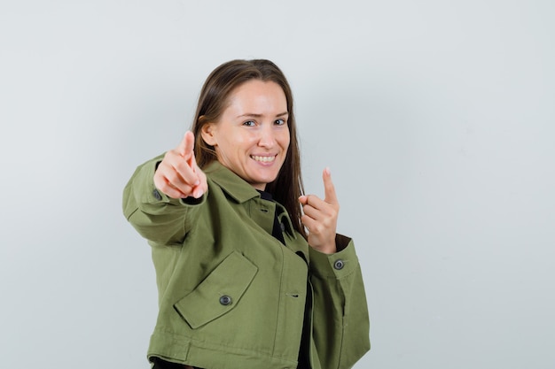 Foto gratuita mujer joven apuntando con chaqueta verde y mirando alegre, vista frontal.