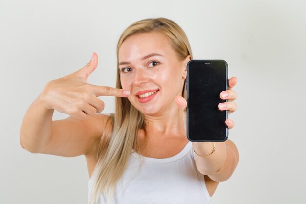 Mujer joven apuntando con el dedo al teléfono inteligente en camiseta y mirando alegre.