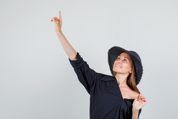 Mujer joven apuntando con el dedo hacia arriba en camisa, sombrero y mirando alegre