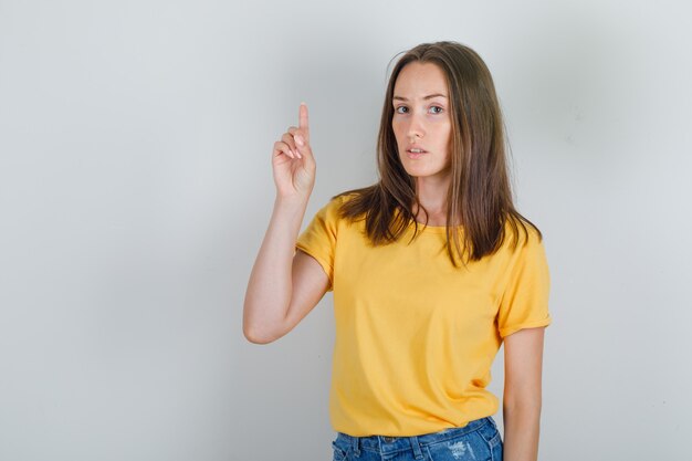 Mujer joven apuntando con el dedo hacia arriba en camiseta amarilla, pantalones cortos y mirando preocupado