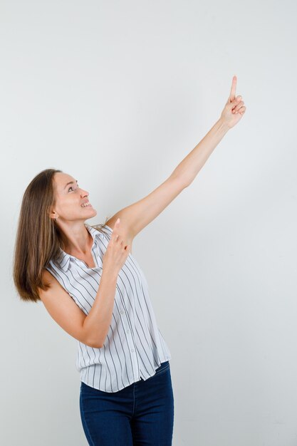 Foto gratuita mujer joven apuntando con el dedo hacia arriba en camiseta, jeans y mirando alegre. vista frontal.