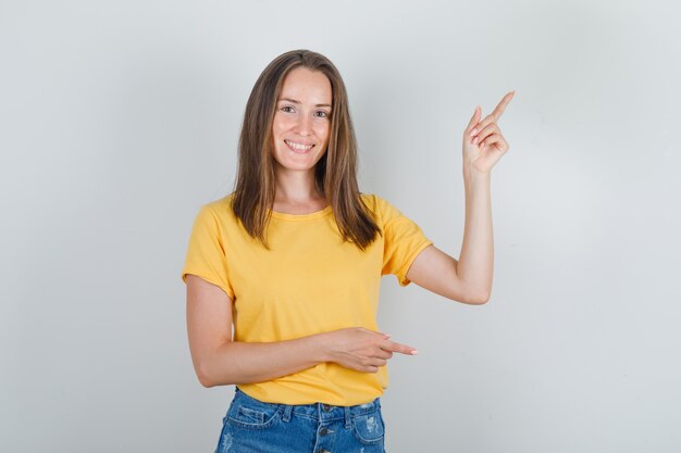 Mujer joven apuntando con el dedo en camiseta amarilla, pantalones cortos y mirando alegre
