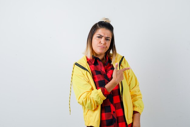 Mujer joven apuntando a la derecha en camisa a cuadros, chaqueta y mirando pensativo. vista frontal.