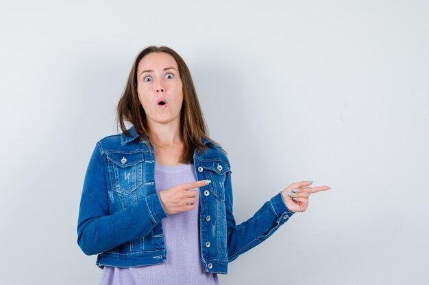 Mujer joven apuntando a la derecha en camiseta, chaqueta y mirando sorprendido, vista frontal.