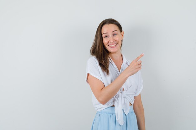 Mujer joven apuntando a la derecha con el dedo índice en blusa blanca y falda azul claro y mirando alegre