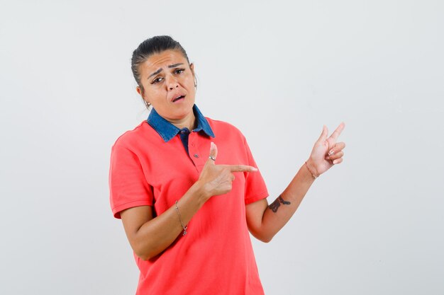 Mujer joven apuntando a la derecha con los dedos índices en camiseta roja y mirando molesto, vista frontal.