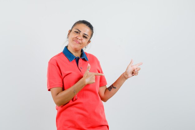 Mujer joven apuntando hacia la derecha con los dedos índices, sonriendo en camiseta roja y luciendo bonita, vista frontal.