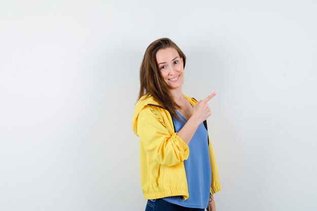 Mujer joven apuntando a la esquina superior derecha en camiseta, chaqueta y mirando alegre, vista frontal.