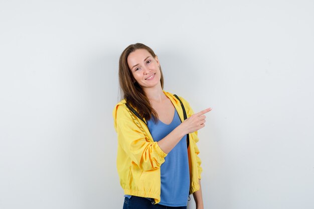 Mujer joven apuntando a la esquina superior derecha en camiseta, chaqueta y mirando confiado, vista frontal.