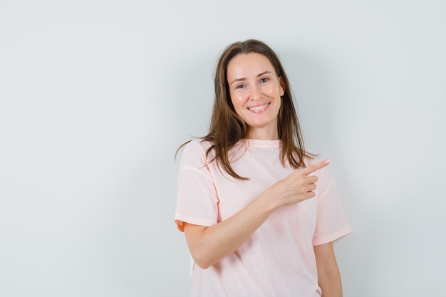 Mujer joven apuntando a la esquina superior derecha en camiseta rosa y mirando feliz. vista frontal.