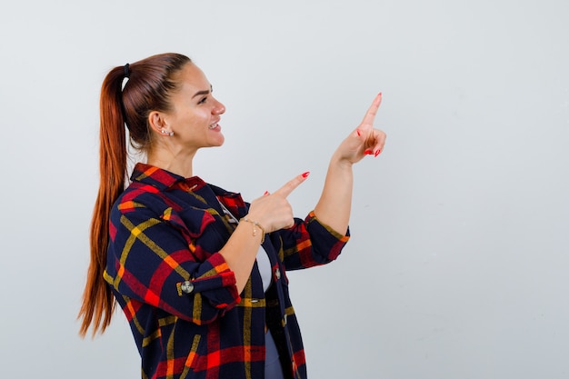 Mujer joven apuntando a la esquina superior derecha en la parte superior de la cosecha, camisa a cuadros y mirando feliz, vista frontal.