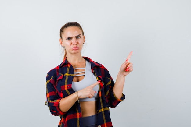 Mujer joven apuntando a la esquina superior derecha en la parte superior de la cosecha, camisa a cuadros, pantalones y mirando ofendido, vista frontal.