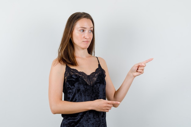 Mujer joven apuntando hacia fuera en camiseta negra y mirando indeciso